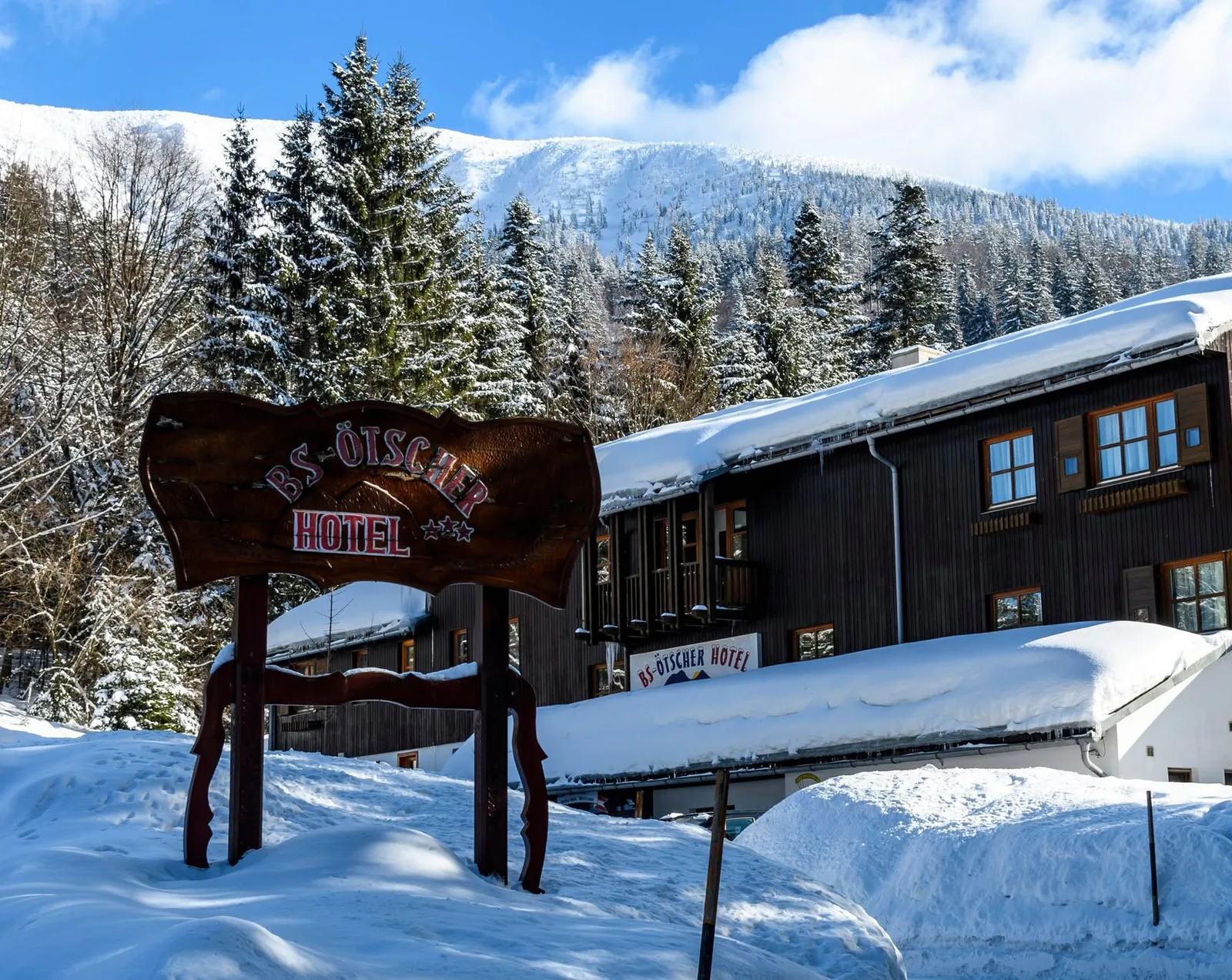 A BS-Ötscher Hotel fa cégtáblája a havas fenyvesekkel és az Ötscherrel a háttérben